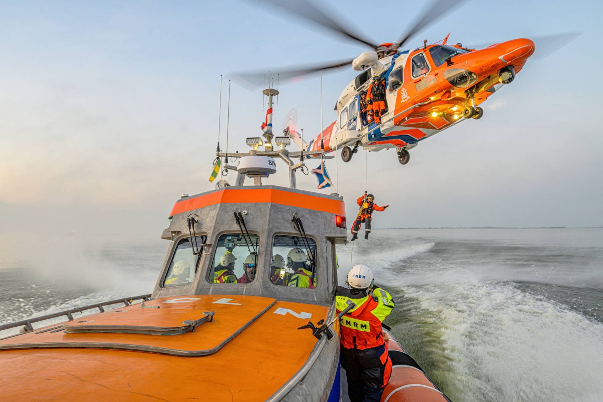 KNRM-räddningsbåt till sjöss med en räddningshelikopter som vinschar upp en besättningsmedlem